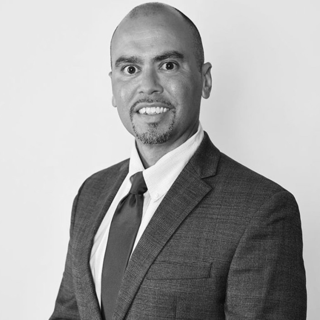 A professionally dressed individual in a suit and tie smiles warmly against a plain background.