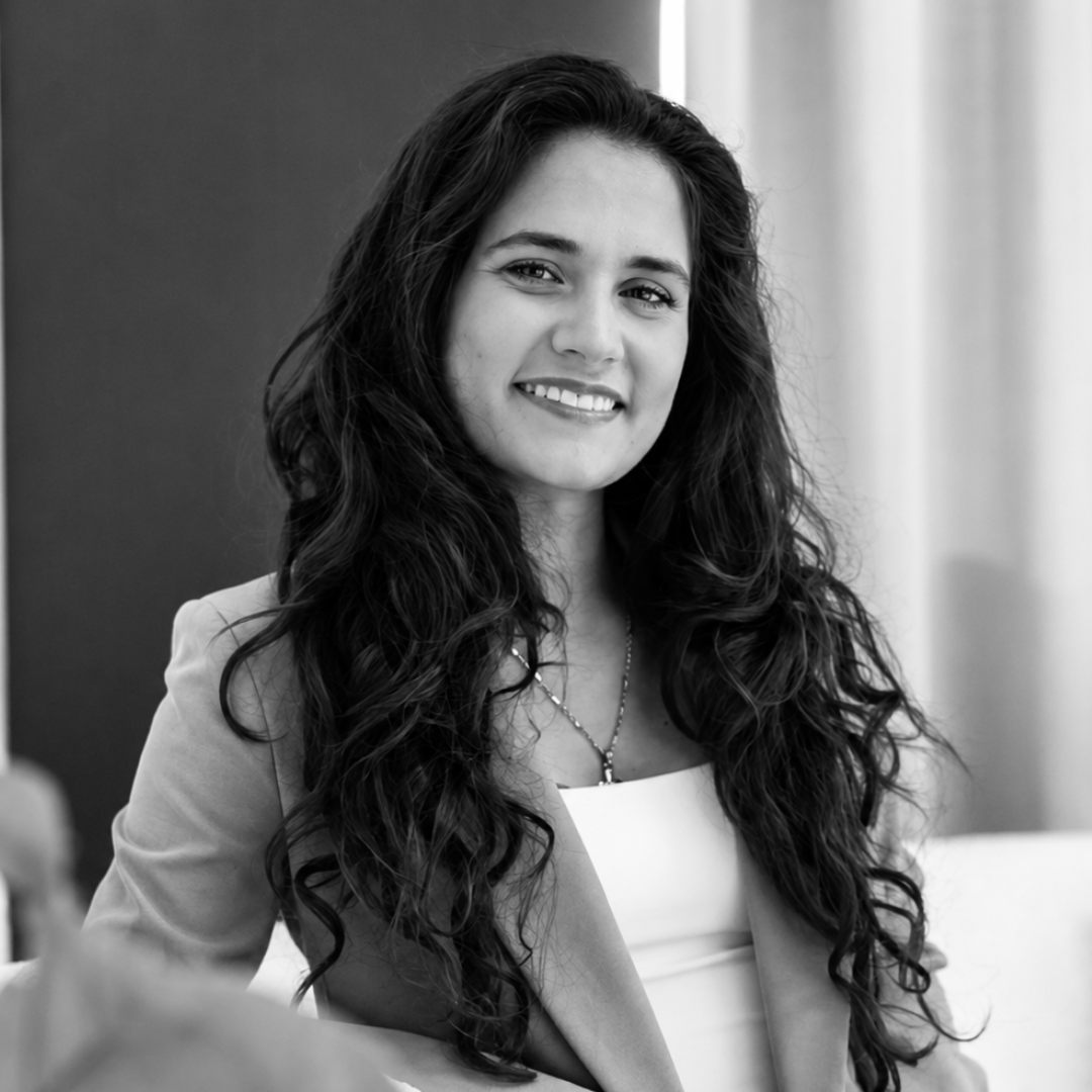 A smiling person with long, wavy hair wears a blazer and necklace, captured in a black-and-white portrait.